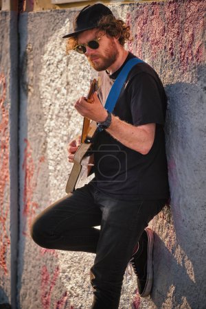 Téléchargez les photos : Musicien de rue hipster en noir jouant de la guitare électrique dans la rue au coucher du soleil appuyé sur un mur. Lisbonne, Portugal - en image libre de droit