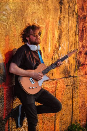 Foto de Hipster músico callejero de negro tocando la guitarra eléctrica en la calle al atardecer apoyado en una pared pintada - Imagen libre de derechos