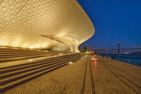 Photo for Lisbon, Portugal - August 08, 2022: MAAT - Museum of Art, Architecture and Technology is a modern building on bank of Tagus river with 25th of April bridge in background in evening. Lisbon, Portugal - Royalty Free Image