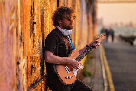 Téléchargez les photos : Musicien de rue hipster en noir jouant de la guitare électrique dans la rue au coucher du soleil appuyé sur un mur peint - en image libre de droit
