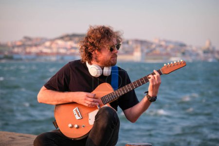 Foto de Hipster músico callejero de negro tocando la guitarra eléctrica en la calle al atardecer en terraplén con Lisboa en el fondo. Portugal - Imagen libre de derechos