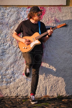 Téléchargez les photos : Musicien de rue hipster en noir jouant de la guitare électrique dans la rue au coucher du soleil appuyé sur un mur - en image libre de droit
