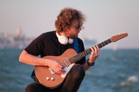 Téléchargez les photos : Musicien de rue Hipster en noir jouant de la guitare électrique dans la rue au coucher du soleil sur un remblai - en image libre de droit