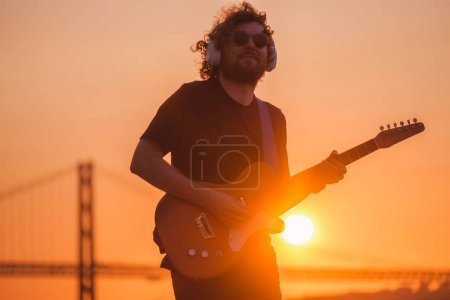 Téléchargez les photos : Musicien de rue Hipster en noir jouant de la guitare électrique dans la rue à l'extérieur au coucher du soleil - en image libre de droit