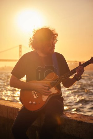 Téléchargez les photos : Musicien de rue Hipster en noir jouant de la guitare électrique dans la rue au coucher du soleil - en image libre de droit
