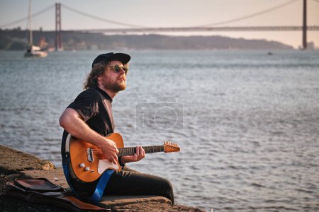 Téléchargez les photos : Musicien de rue Hipster en noir jouant de la guitare électrique dans la rue assis sur le quai au coucher du soleil - en image libre de droit