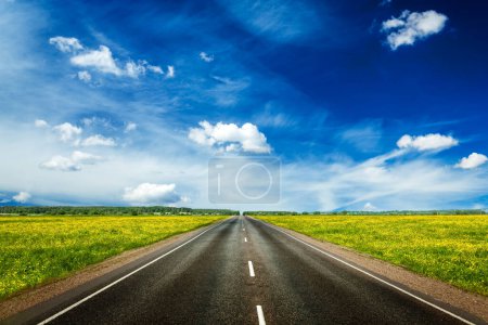 Téléchargez les photos : Concept de voyage arrière-plan - une route vide avec un ciel bleu et des champs de printemps verts en fleurs de chaque côté - en image libre de droit