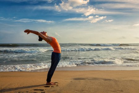 Foto de Mujer joven haciendo yoga Saludo al sol Surya Namaskar pose Hasta Uttanasana en la playa tropical al atardecer - Imagen libre de derechos