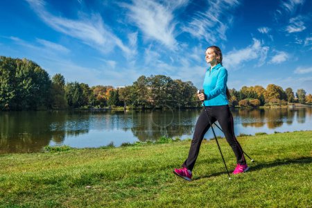 Foto de Aventura a pie nórdica y ejercicio - mujer joven haciendo senderismo con bastones de senderismo nórdicos en el parque a lo largo del río - Imagen libre de derechos