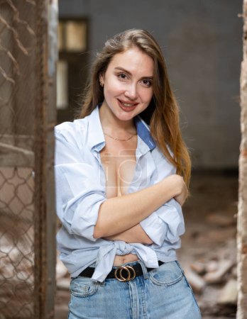Attractive brown-haired young woman in blue men shirt, tied in knot on tummy, slightly revealing breasts, posing near old rusty wire fencing in dilapidated abandoned house