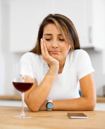 Upset young woman with glass of red wine in kitchen reading messages on mobile phone screen