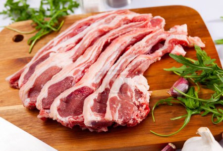 Raw hogget chops on ribs on wooden board with fresh fragrant arugula leaves, garlic and seasonings. Main ingredients for cooking