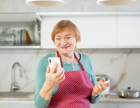 Cheerful senior woman searching information on internet using phone in her kitchen