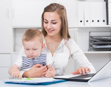 Satisfied female with kid is productively working behind laptop in office.