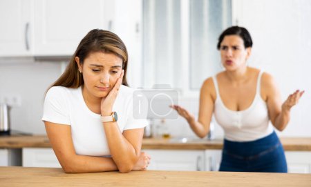Unhappy offended young Latina standing in kitchen at home, listening to her disgruntled female friend. Concept of problems in friendships