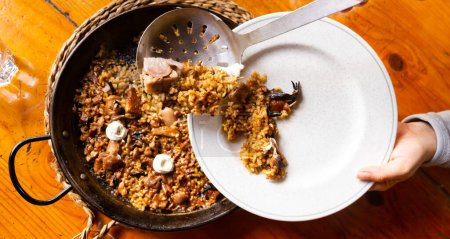 Customer pouring delicious Catalan mountain rice with mushrooms and pork feet into plate from traditional paella pan in local restaurant