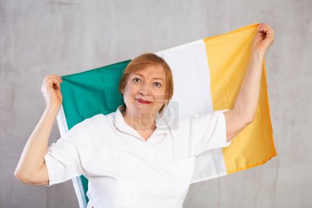 Smiling patriotic elderly woman waving national Irish tricolor flag while posing against gray studio background