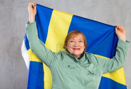 Portrait of smiling elderly woman holding in hands national flag of Sweden, posing against gray studio background