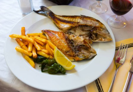 Fried cut dorada fish served on white plate with vegetable garnish and lemon..