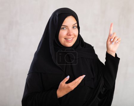 Mujer joven en hiyab negro señalándote - Retrato de mujer joven señalando con el dedo. Aislado sobre fondo gris