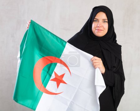 Eine ruhige Muslimin im traditionellen schwarzen Hidschab hält die algerische Flagge in der Hand. Porträt einer Muslimin mit algerischer Flagge auf grauem Hintergrund