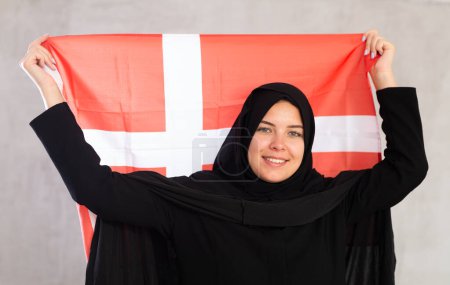 tranquila mujer musulmana sonriente en hiyab negro tradicional sostiene la bandera de Dinamarca. Retrato de mujer musulmana con bandera danesa sobre fondo gris