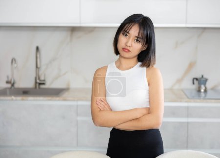 Upset young Asian woman standing in melancholic pose against background of white kitchen cabinet