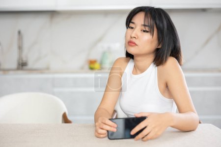 Empörte junge Asiatin sitzt in melancholischer Pose und hält Handy in der Küche