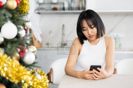 Sad young Asian girl waiting for phone call sitting at table by Xmas tree in the kitchen