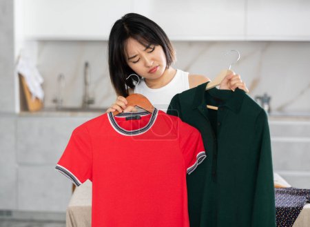 Verärgerte junge Asiatin schaut unbefriedigend auf Kleider, die zur Auswahl in der Hand gehalten werden