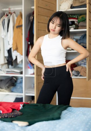 Trauriges junges asiatisches Mädchen versucht, ein Kleid zu wählen, das neben einem Kleiderschrank mit offenen Türen steht