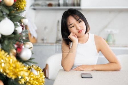 Verärgerte junge Asiatin sitzt traurig mit Handy in der mit Weihnachtsbaum geschmückten Küche