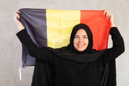 sonriente mujer musulmana en hiyab negro tradicional sostiene la bandera de Bélgica. Retrato de mujer musulmana con bandera belga sobre fondo gris