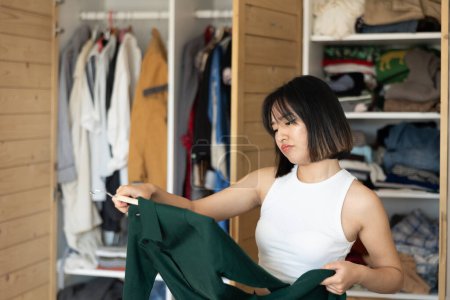 Upset young Asian woman looking at dresses held in hand for choice in dressing room