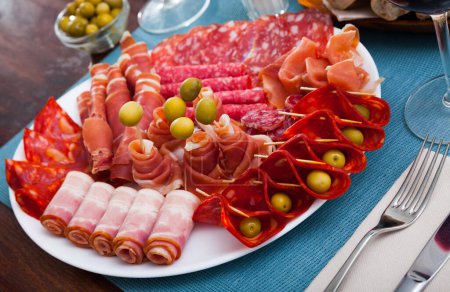 Slices of Spanish dry-cured gammon, variety of sausages and bacon served with green olives on round plate..