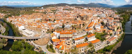 Blick von der Drohne auf die Wohngegend der Stadt Plasencia mit bräunlich gefliesten Häuserdächern und der römisch-katholischen Kathedrale am grünen Ufer des mäandernden Flusses Jerte im Frühling, Caceres, Spanien