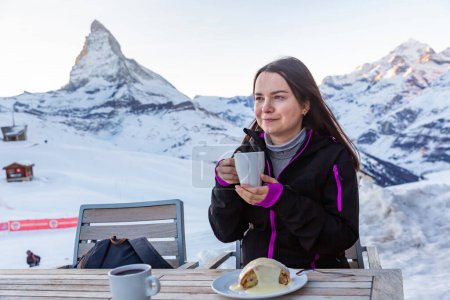 Smiling woman enjoying cup of hot mulled wine with delicious strudel in outdoor cafe of ski resort against backdrop 