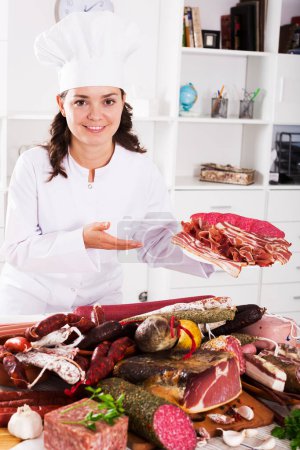 Cocinar mujer cuesta cerca de la mesa con productos ahumados y salchichas y sostiene en placa de mano con jamón