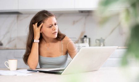 Young puzzled woman working remotely on laptop at table in kitchen at home