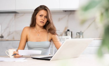 Young puzzled woman working remotely on laptop at table in kitchen at home