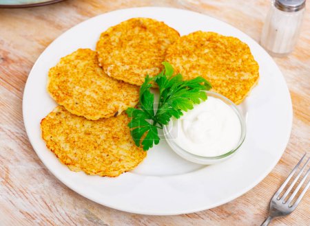 Fried potato pancakes latke are neatly laid out on plate and decorated with parsley leaves. Dish is complemented with fresh sour cream and cup of hot herbal tea.