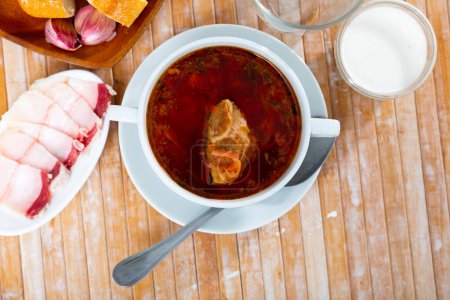 Portion of national Ukrainian dish borscht served in bowl on table with sliced salo.