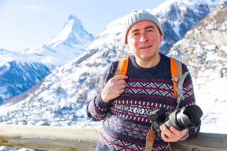 Positive adult man with passion for photography traveling in Swiss Alps, standing with camera on background of snowy mountain range with sharp Matterhorn peak on sunny winter day..