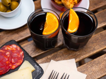 Vermú rojo dulce aromatizado adornado con rodajas de naranja servidas en dos vasos con chorizo, queso y aceitunas. Hora tradicional del vermut español