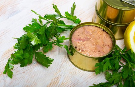 Closeup view of opened can of tuna in oil on wooden table with lemon and greens