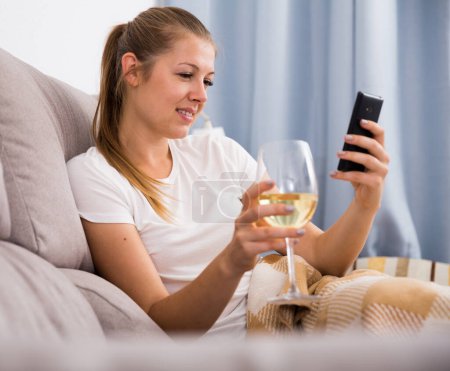 Young smiling female with modern mobile phone and wine on sofa indoors