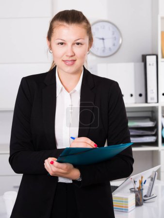Mujer en traje negro está firmando papeles de acuerdo de naturaleza financiera en la oficina.