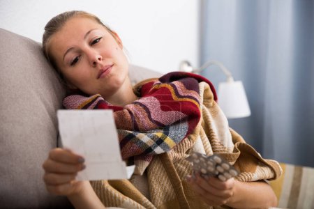 Sad young female lying at sofa reading recipe with pills