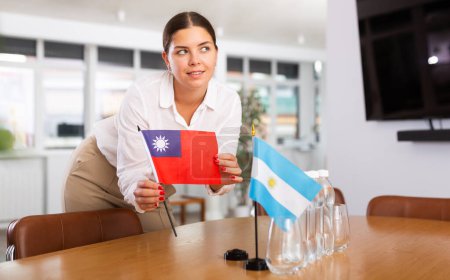 Jeune femme positive mettant petit drapeau de Taiwan sur la table à côté du drapeau de l'Argentine et des bouteilles d'eau dans la salle de conférence