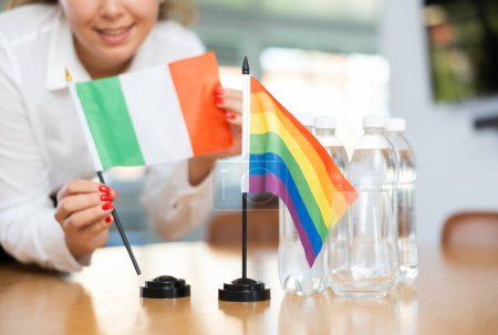 Unrecognizable woman preparing room for international negotiations and communication discussions of leaders. Lady sets miniatures flags of LGBT and Italy on table. Unfocused shot